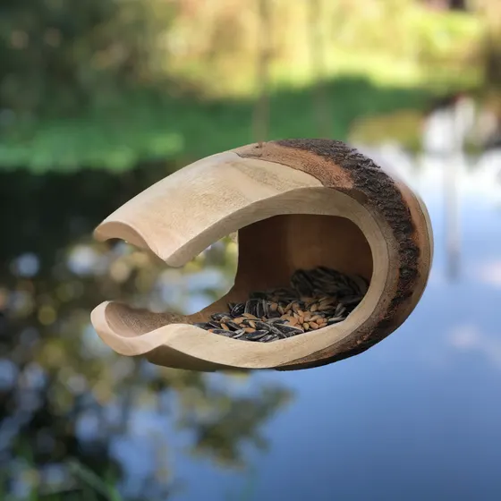Une mangeoire en bois de manguier, ouverte et garnie de graines de tournesol, suspendue au-dessus d’un plan d’eau.